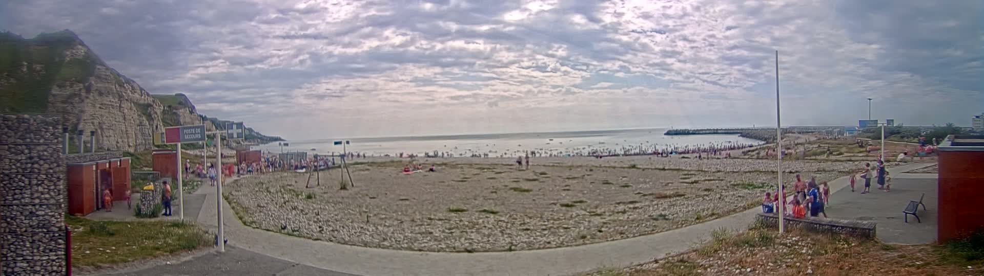 A pebble beach with many people on it and in the water, under a partly cloudy sky, is situated below a tall, light-colored cliff.