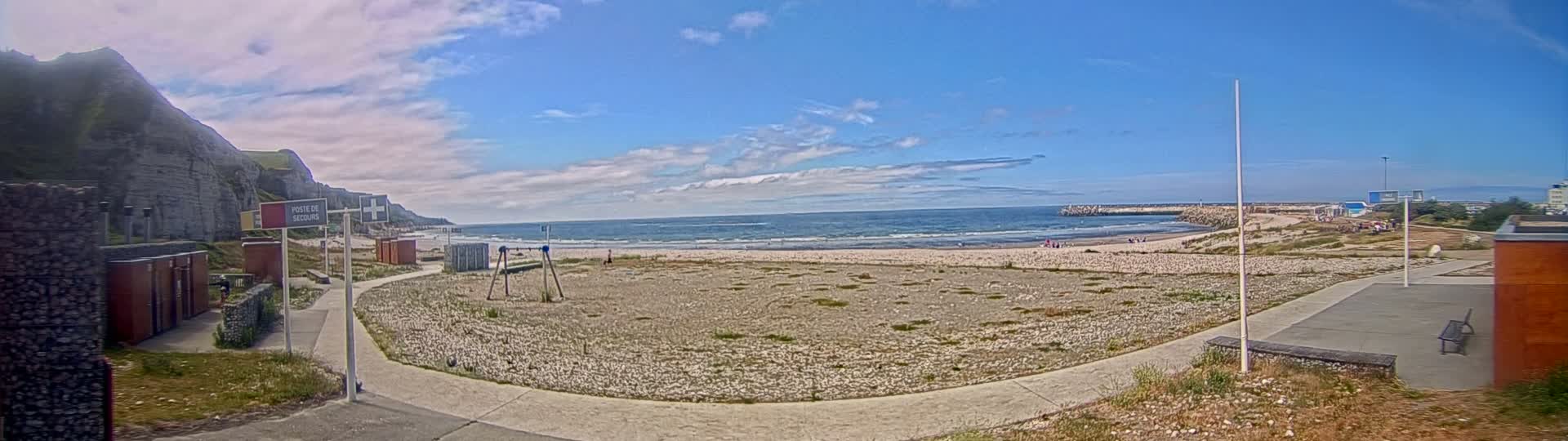 A mostly sunny day shows a sparsely populated beach with a rocky cliff in the background, a calm ocean, and a paved pathway leading to small buildings near the beach.