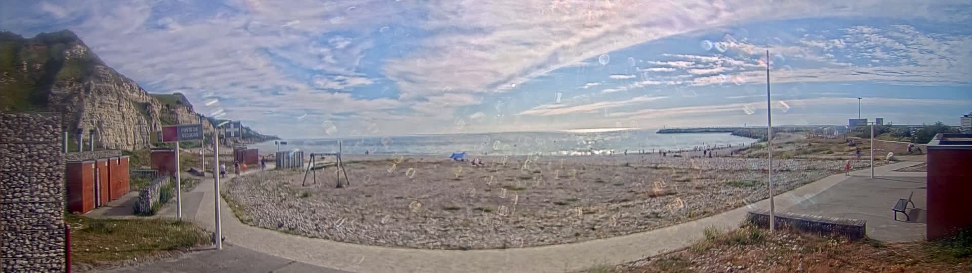 A pebble beach with a few people on it sits below a tall, light-colored cliff face under a mostly sunny sky with scattered clouds.