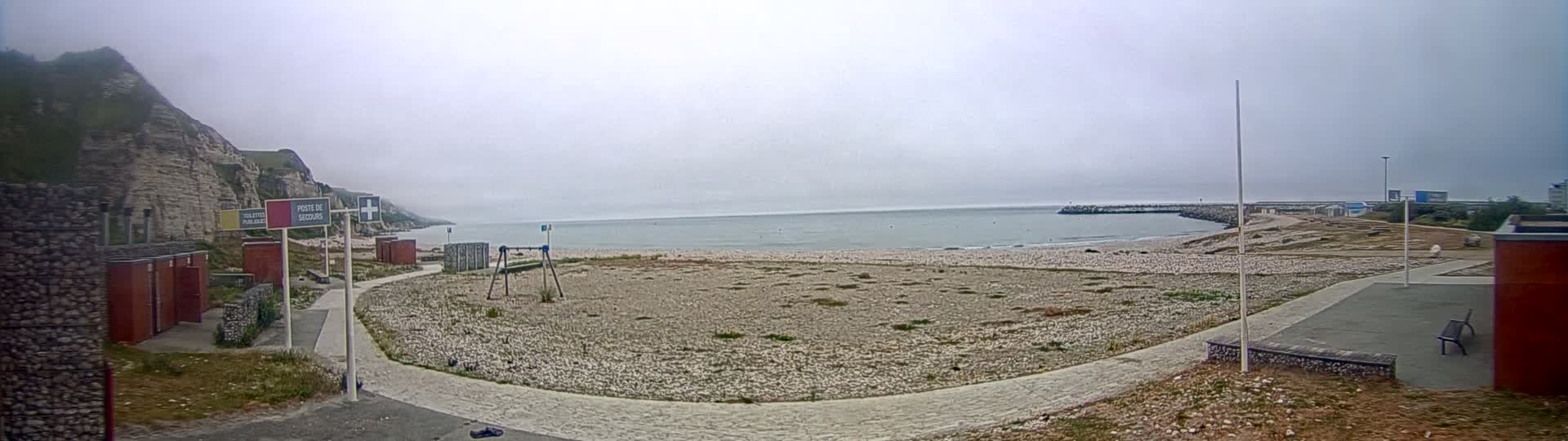 A mostly empty pebble beach stretches between a rocky cliff and a breakwater on an overcast day.