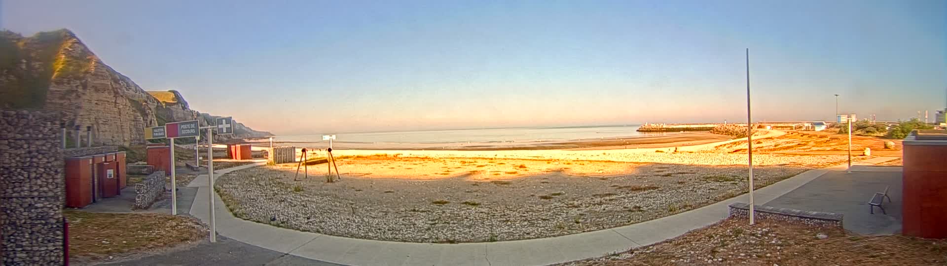 A sandy beach stretches out to a calm sea under a clear, light-blue sky at sunset, with a rocky cliff to the left and a paved walkway leading to the beach.