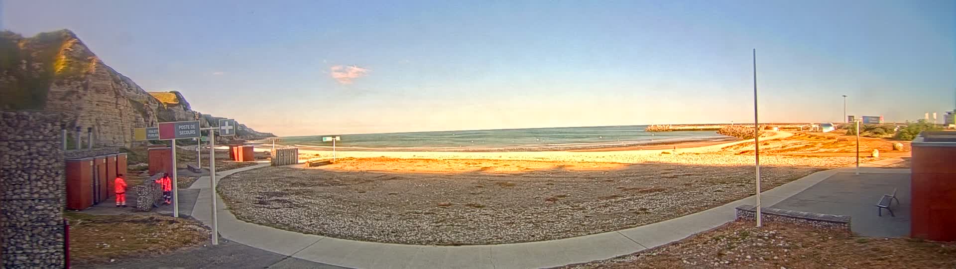 A sandy beach stretches along a calm ocean under a clear, bright sky, with a rocky cliff and paved walkway to the left and a small building to the right.