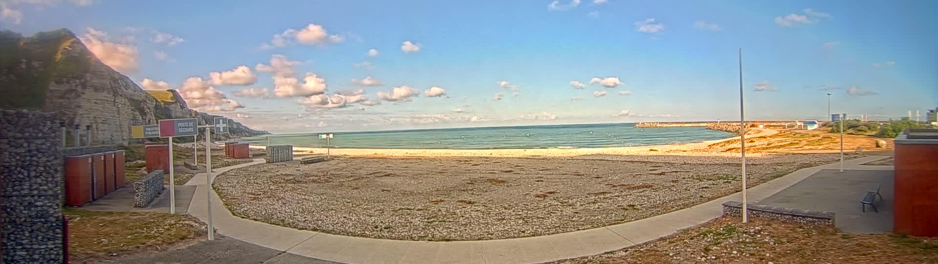 A mostly sunny day at a beach with a calm sea, white sand, and a rocky cliff in the background, featuring a paved pathway leading to a small building.
