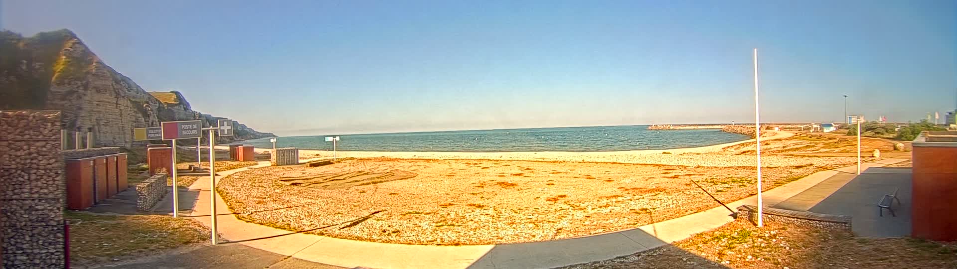 A sandy beach with a calm sea under a clear blue sky is bordered by a rocky cliff on one side and a low wall on the other.