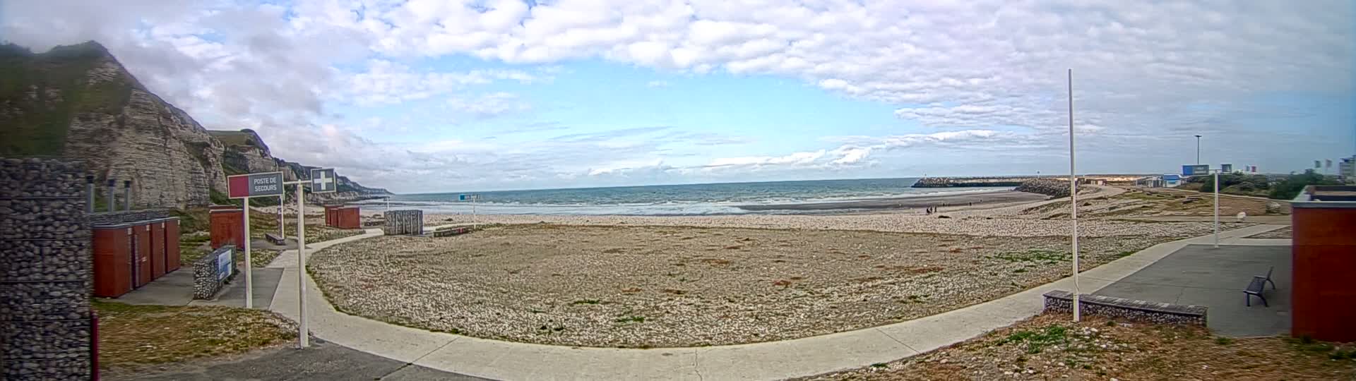 A mostly sunny day shows a beach with a few people, a rocky outcropping, and a paved walkway leading to some small buildings near a cliff.