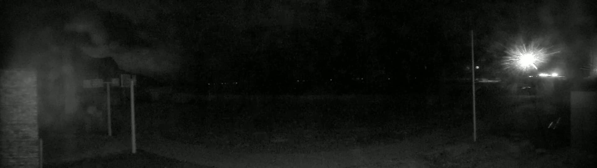 A nighttime, low-light outdoor scene shows a dark area with indistinct objects, a bright light source in the upper right, and faint lights in the distance.
