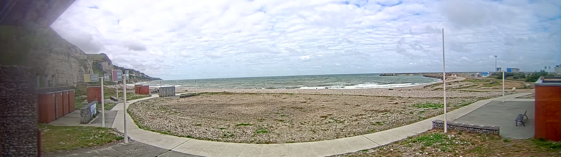 A mostly cloudy day shows a pebble beach with a calm to moderately choppy sea, white cliffs in the background, and a paved walkway leading to some small buildings.