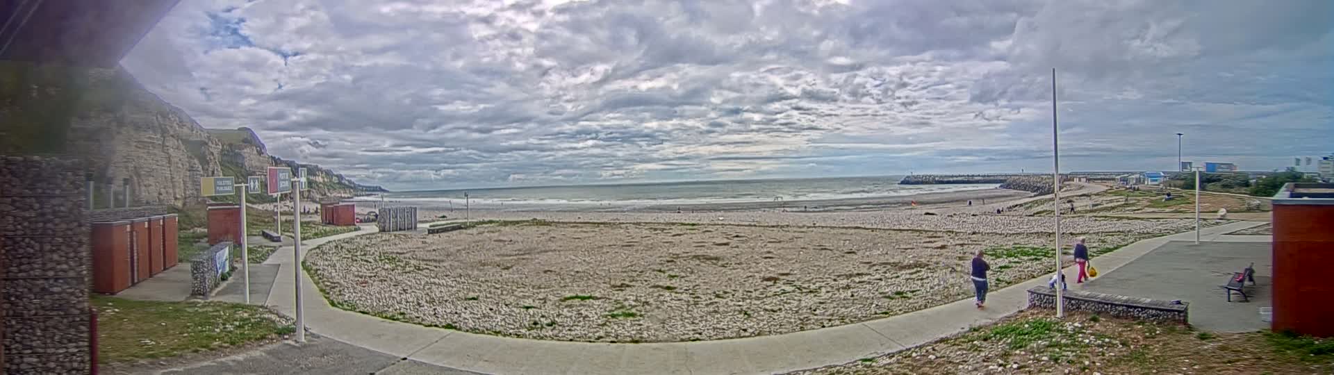A mostly cloudy day shows a pebble beach with a calm to moderately choppy sea, white cliffs in the background, and a paved walkway leading to some small buildings.