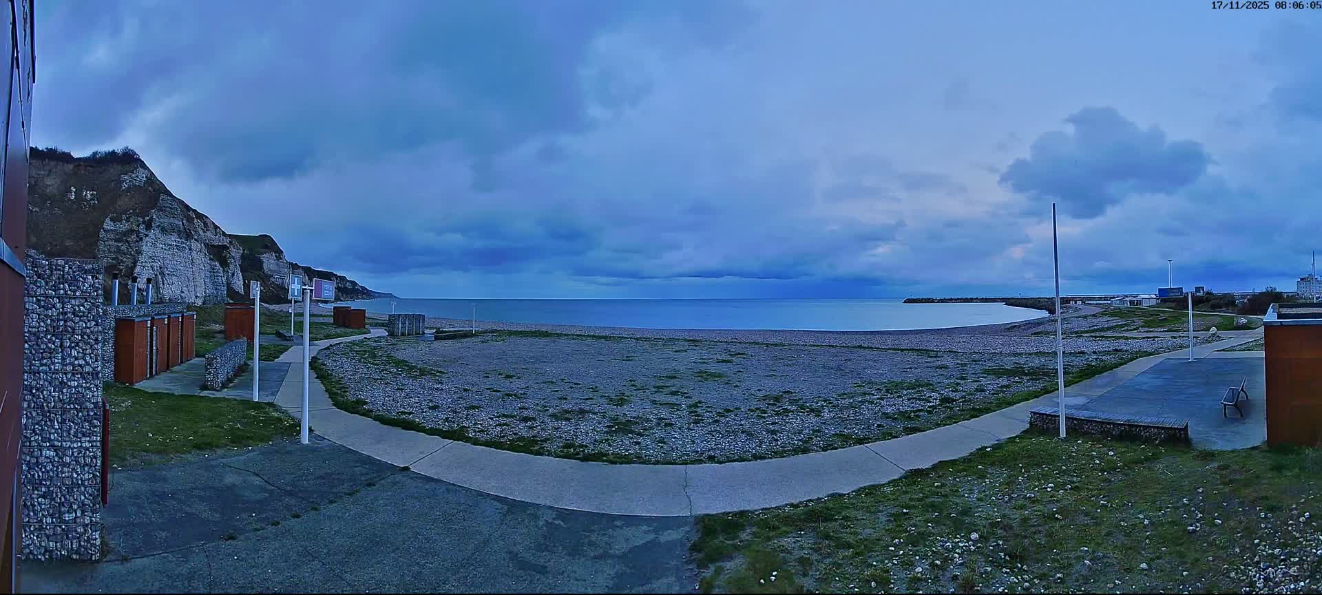 A panoramic view displays a wide pebble beach with white cliffs on the left and several small brown structures, extending towards a calm sea and a distant port under a dim, heavily overcast sky.