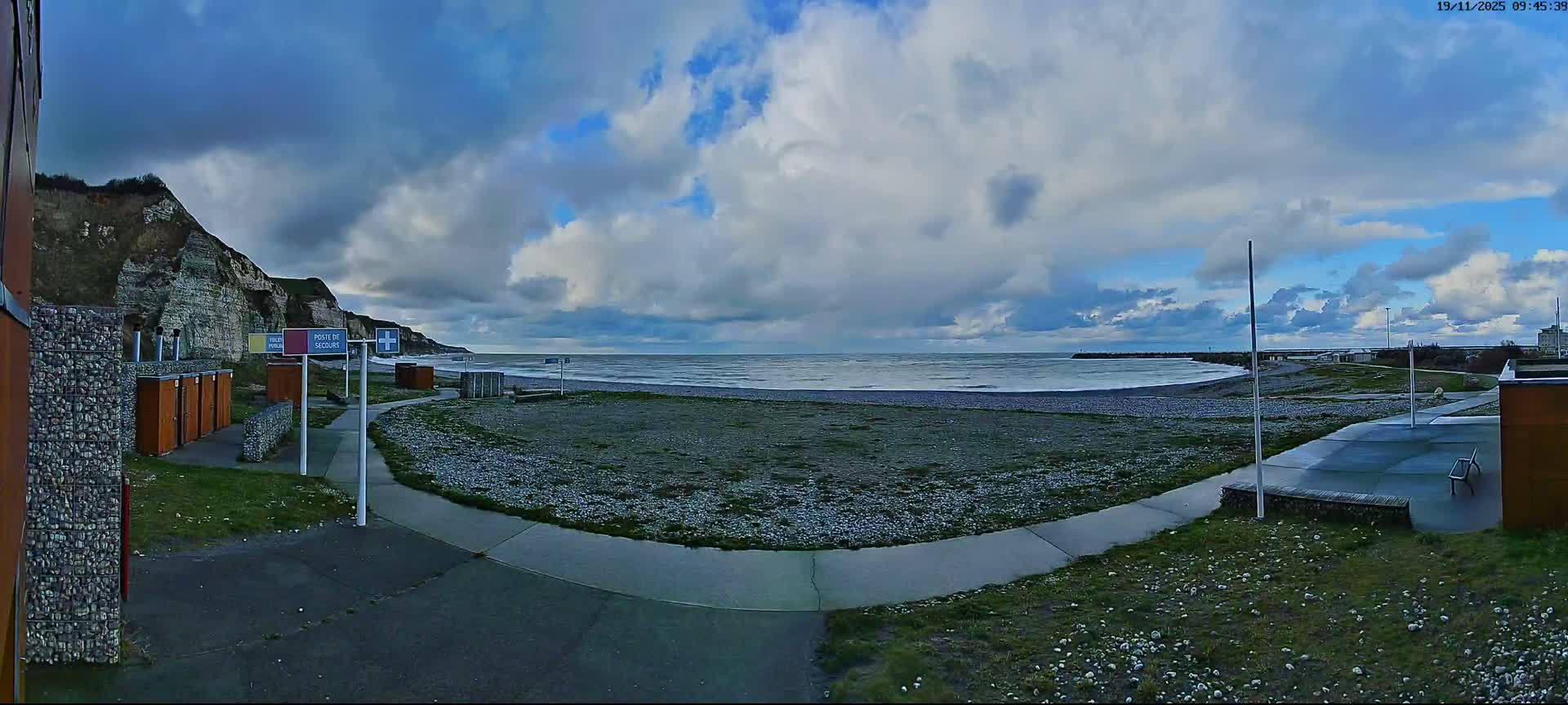 A wide panoramic view captures a pebble beach stretching along a calm sea with cliffs on the left and a distant pier on the right, all under a partly cloudy sky.