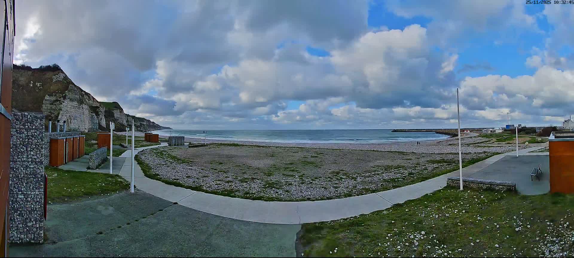 A panoramic view shows a pebbled beach with gentle waves, bordered by white cliffs on the left, a winding pathway, and scattered beach structures under a partly cloudy sky.