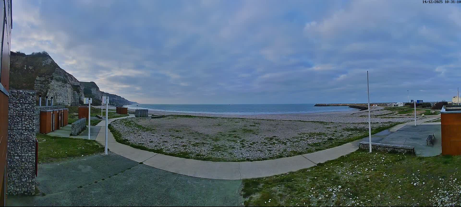 A panoramic view shows a pebbled beach with gentle waves, bordered by white cliffs on the left, a winding pathway, and scattered beach structures under a partly cloudy sky.