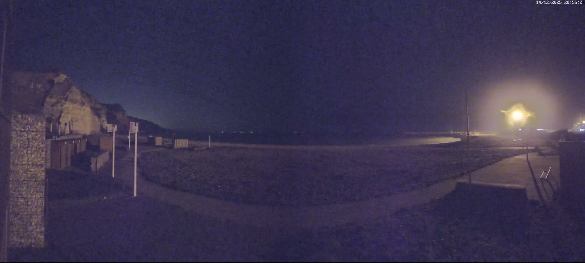 A panoramic view shows a pebbled beach with gentle waves, bordered by white cliffs on the left, a winding pathway, and scattered beach structures under a partly cloudy sky.