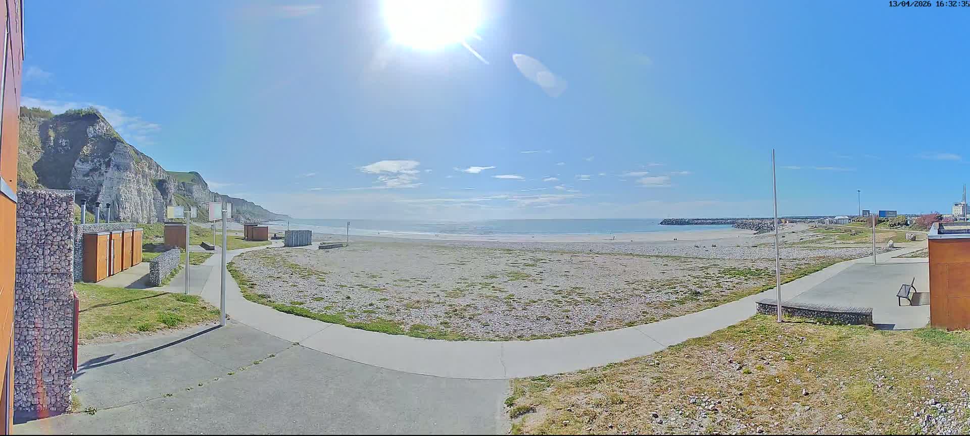 A panoramic view shows a pebbled beach with gentle waves, bordered by white cliffs on the left, a winding pathway, and scattered beach structures under a partly cloudy sky.