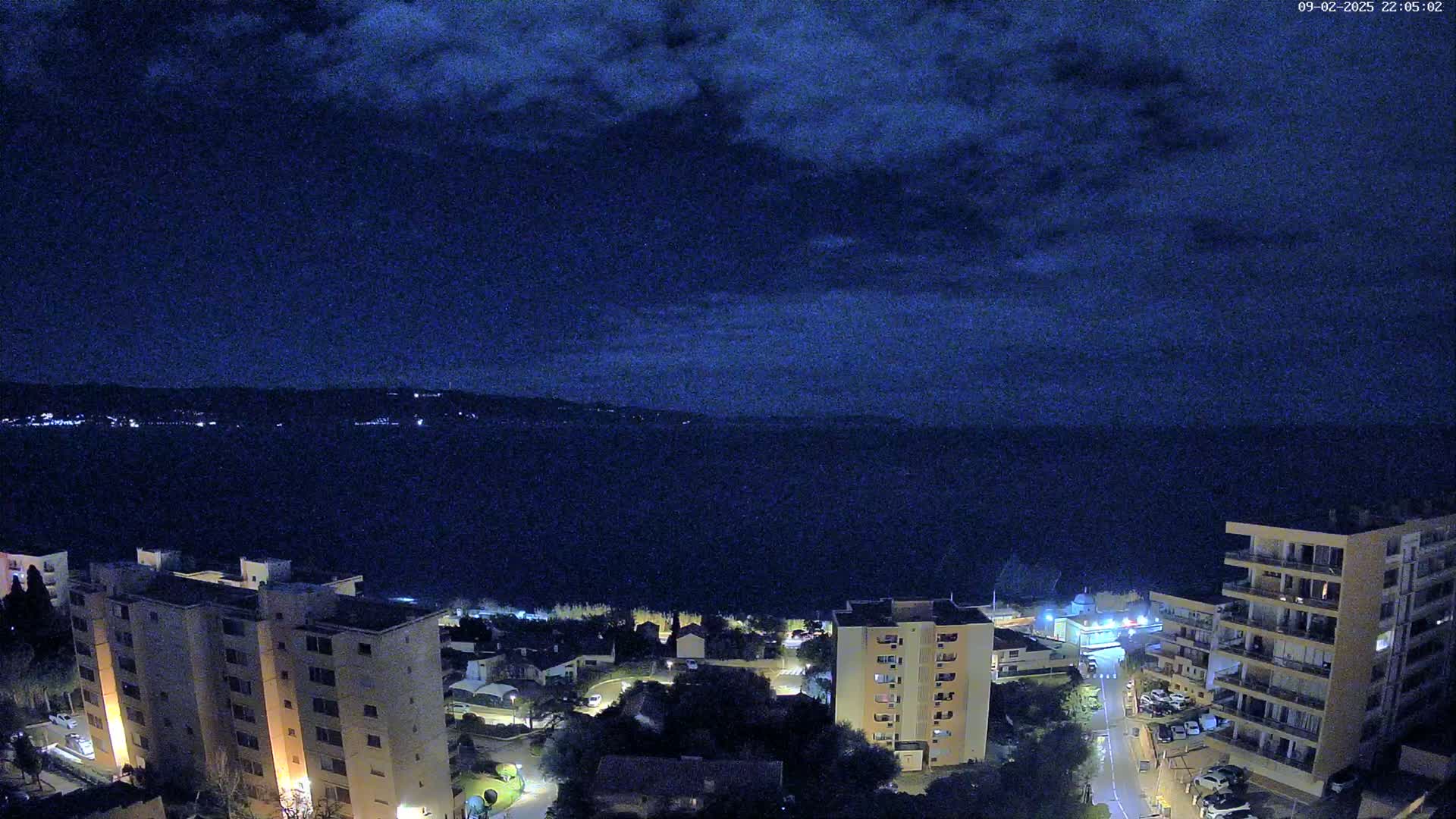 A nighttime view of a coastal town, with dark clouds overhead and a calm body of water extending to a distant, dimly lit shoreline.