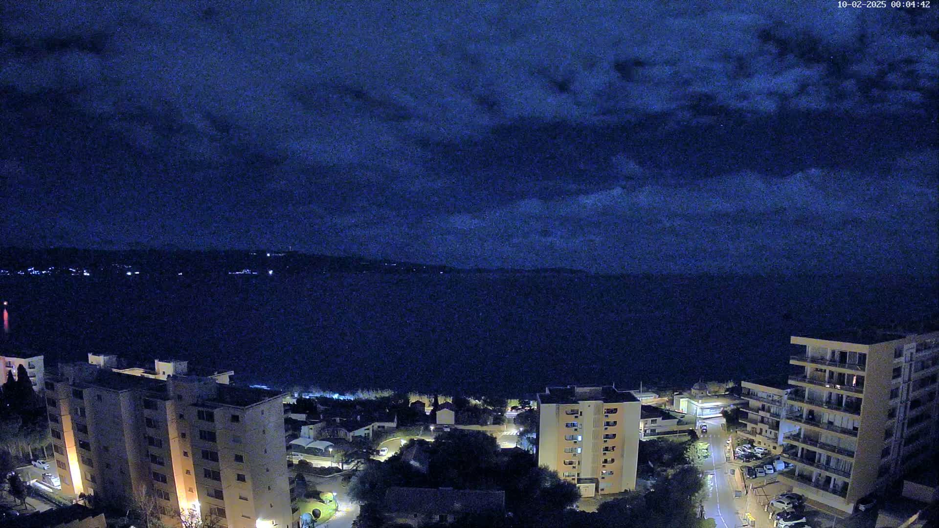 A nighttime view of a coastal town shows several apartment buildings illuminated against a dark, cloudy sky and a calm, dark body of water.