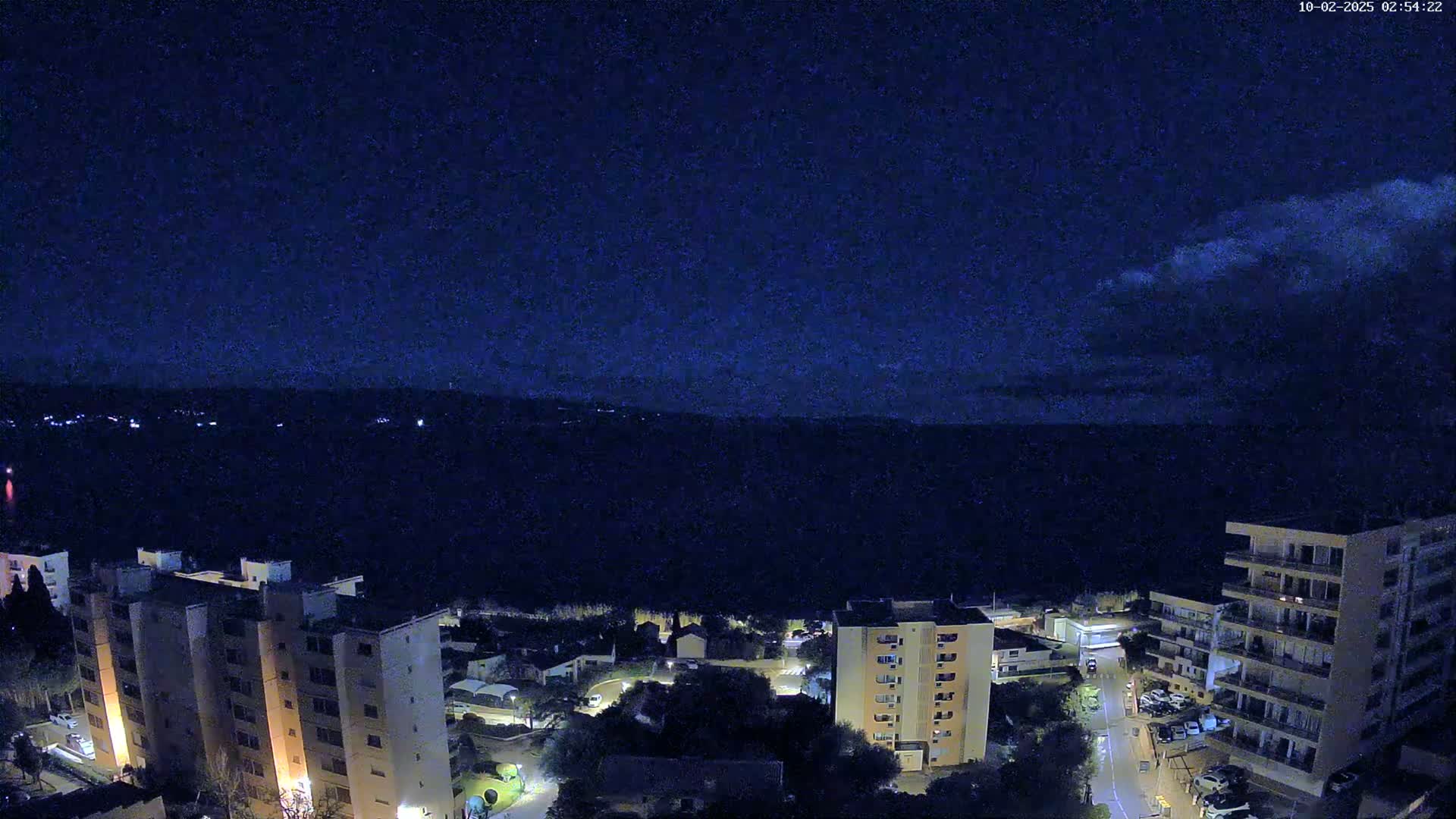 A nighttime view of apartment buildings and a residential area under a mostly clear, dark blue sky with a few high clouds.