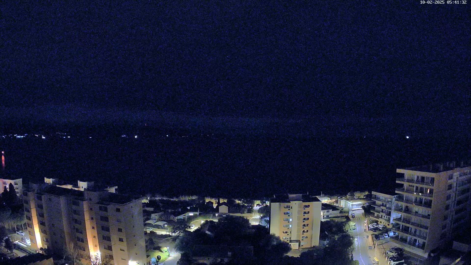 A nighttime, high-angle view shows a dark body of water, a line of lights in the distance, and several multi-story buildings illuminated against a dark sky.