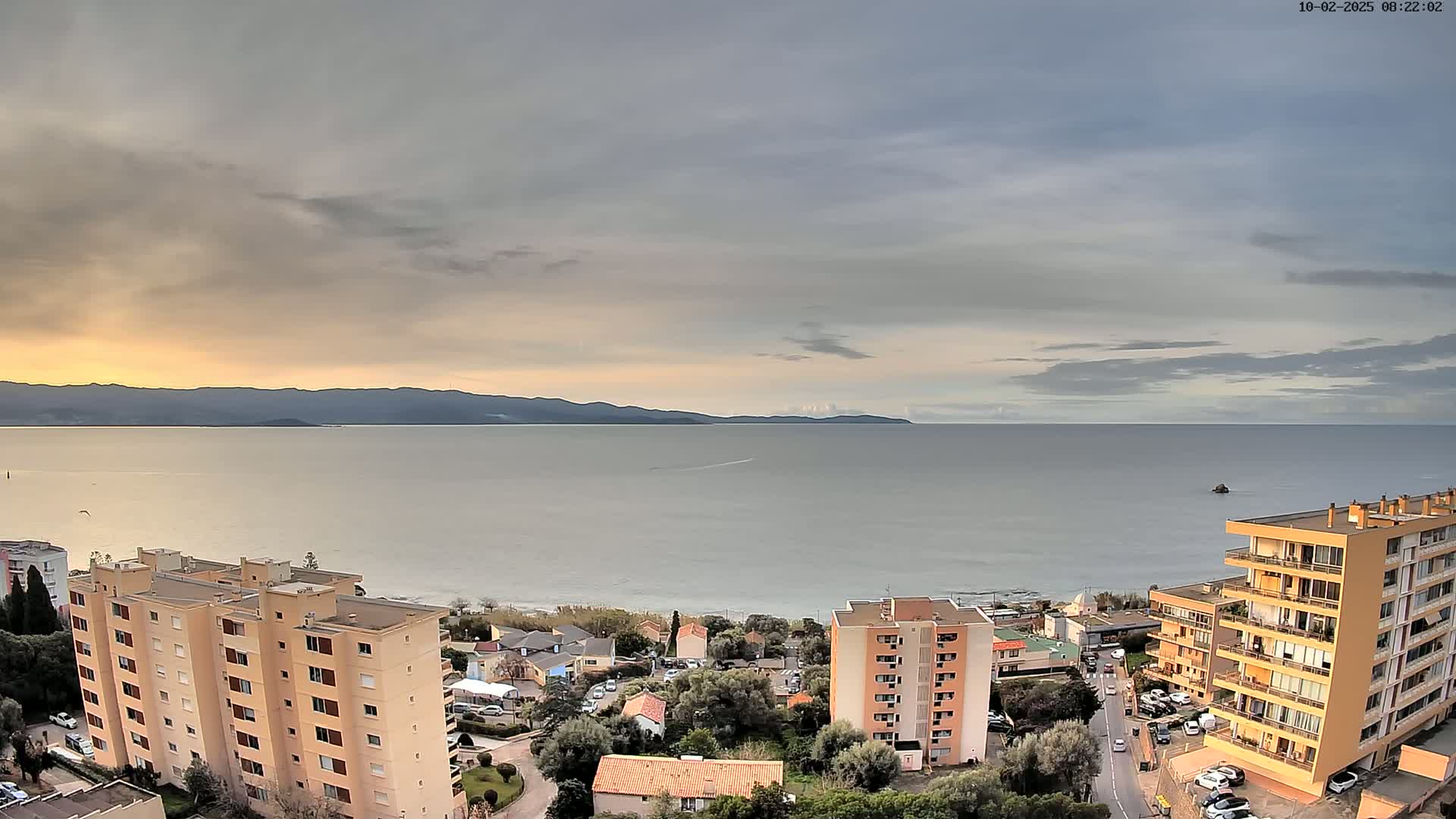 A coastal town with several apartment buildings is visible under a mostly cloudy sky, overlooking a calm sea and distant mountains.