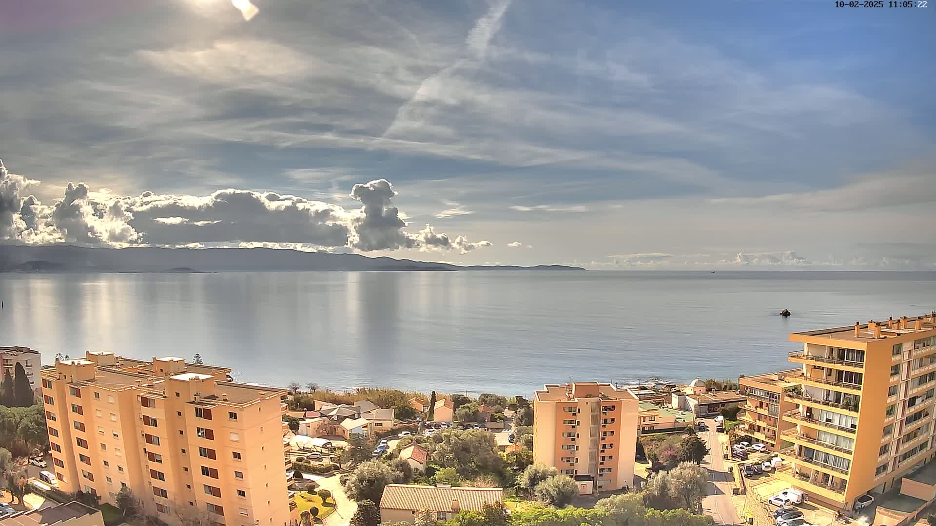 A partly cloudy day overlooks a calm sea with a distant mountain range, and several multi-story buildings are visible in the foreground.