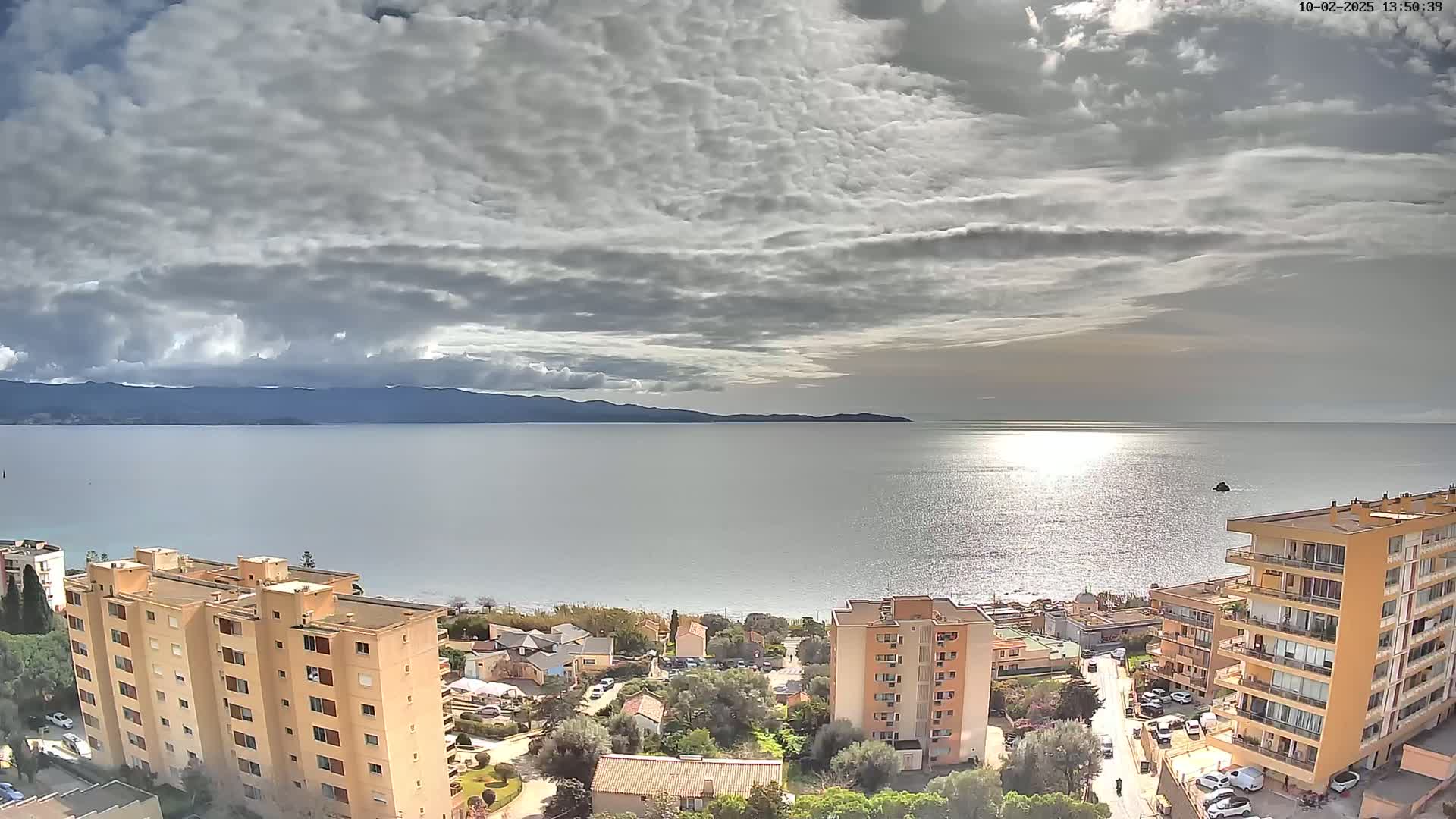 A mostly cloudy sky overlooks a calm sea with a distant mountain range, and several multi-story buildings are visible in the foreground.