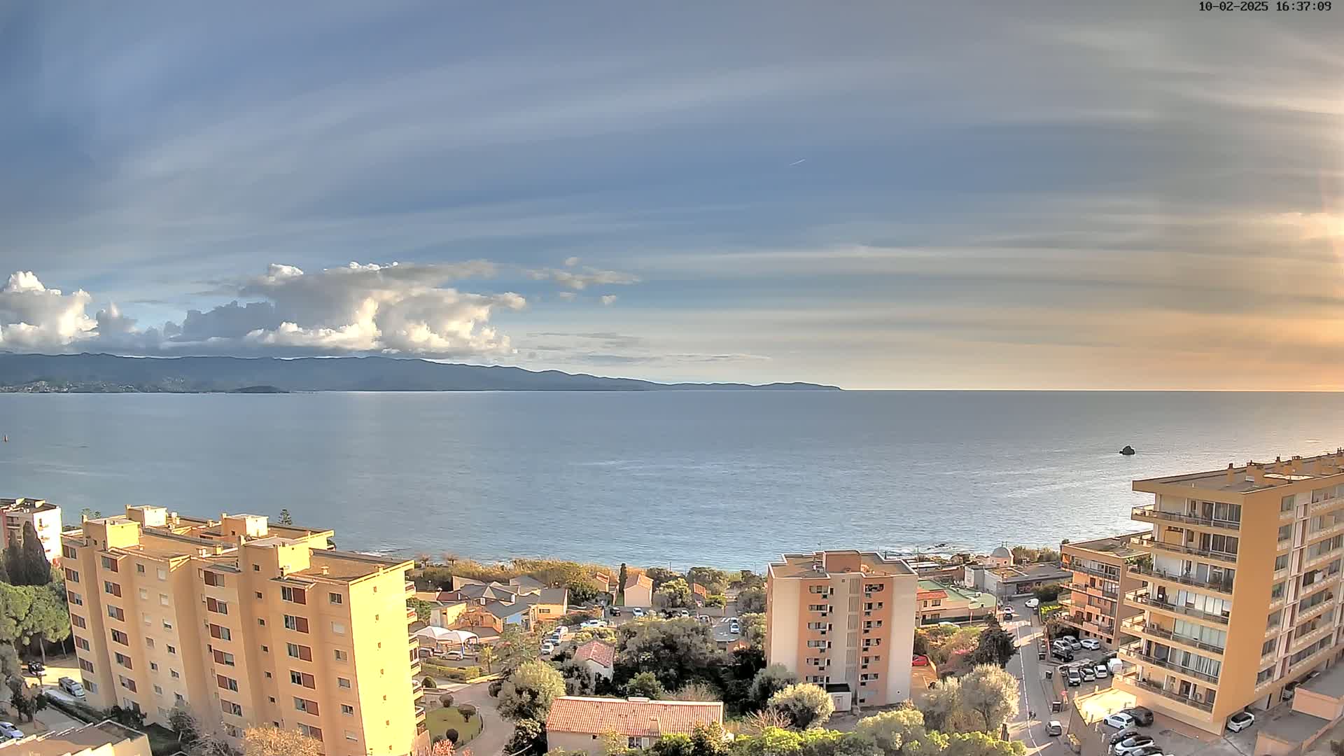 A partly cloudy sky overlooks a calm sea with mountains in the distance, and buildings and trees are visible closer to the foreground.