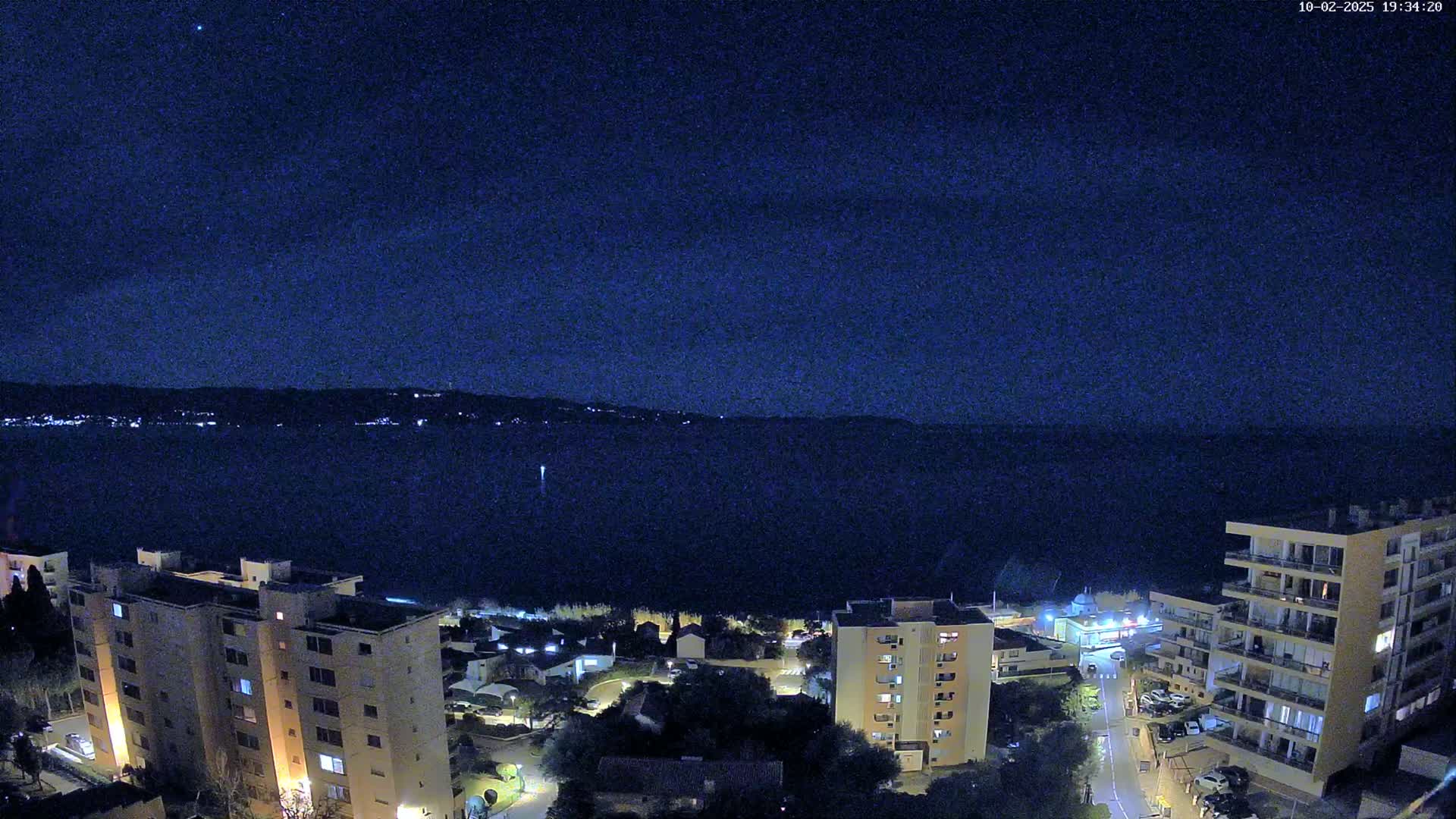 A nighttime view of a calm body of water bordered by apartment buildings and a town under a clear, starry sky.