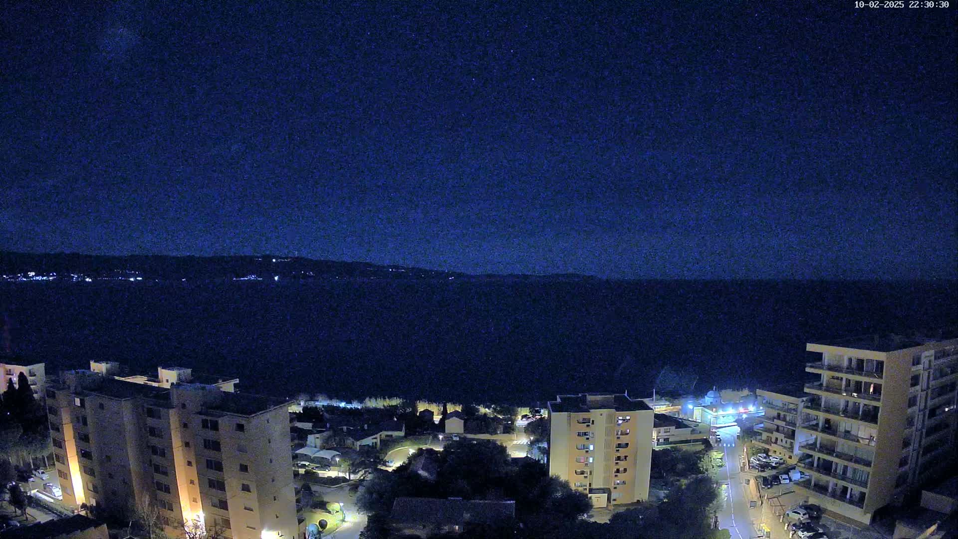 A nighttime view of a coastal town, with apartment buildings and a calm dark body of water under a clear, starry sky.