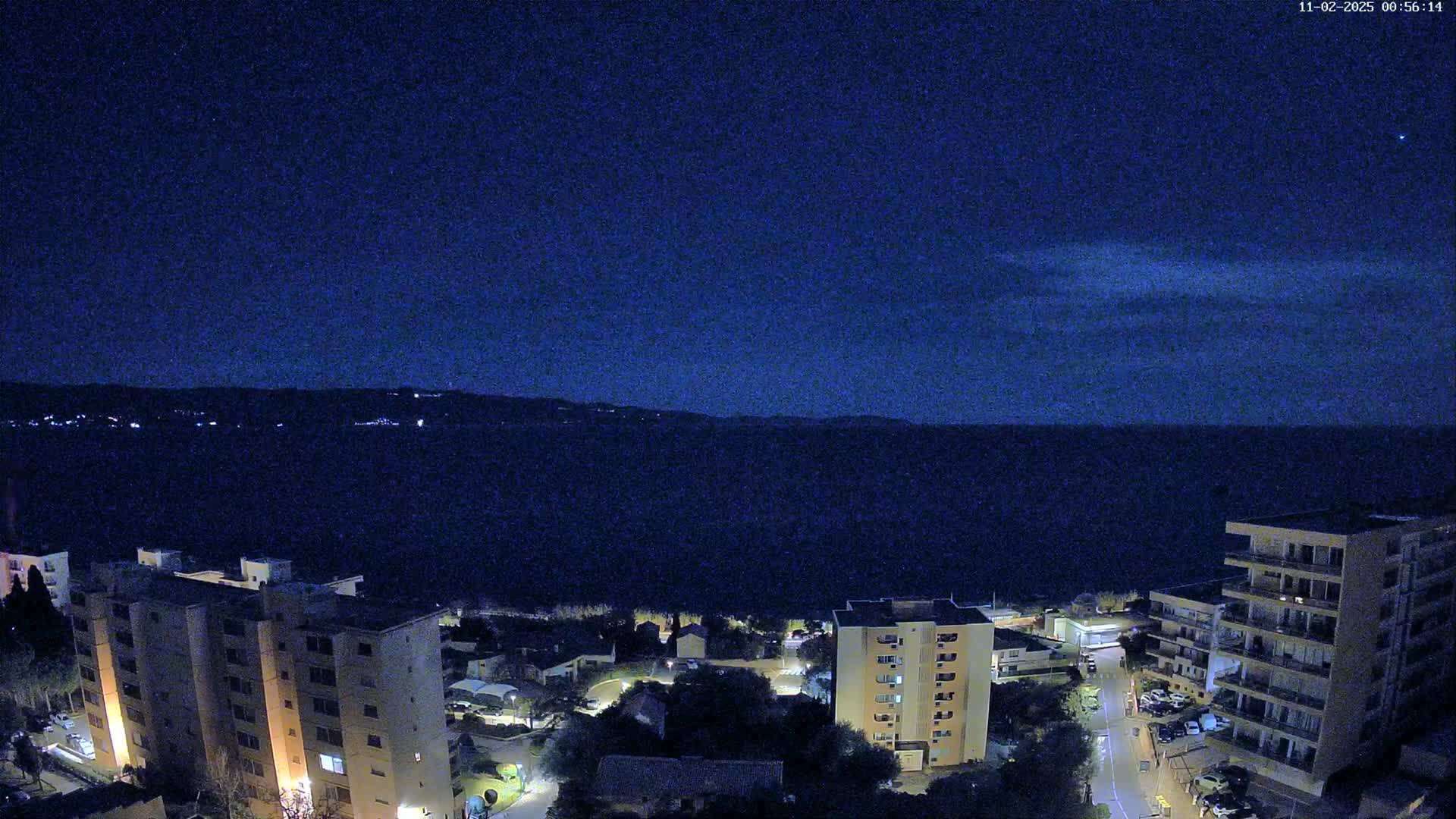 A nighttime aerial view shows a dark body of water under a mostly clear, dark blue sky, with several multi-story buildings and some smaller structures illuminated near the water's edge.
