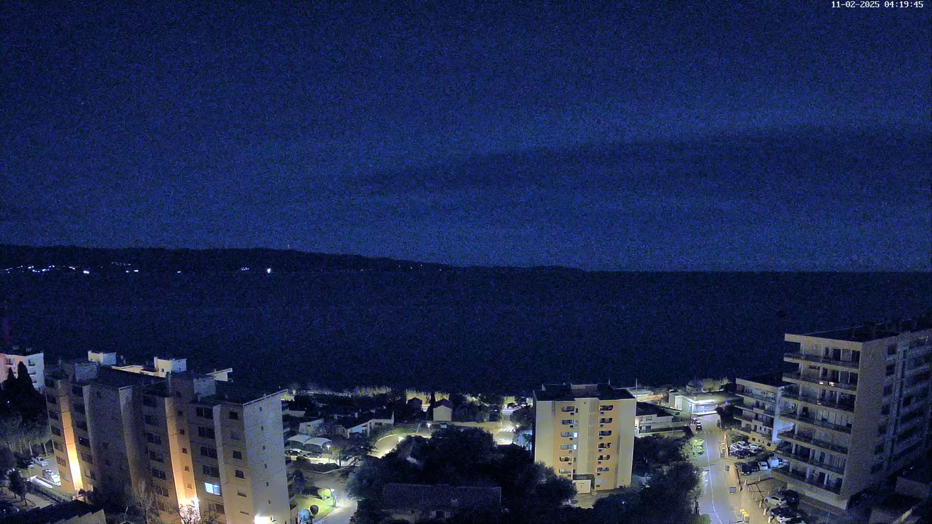 A nighttime, high-angle view shows several apartment buildings near a dark body of water under a clear, dark blue sky.