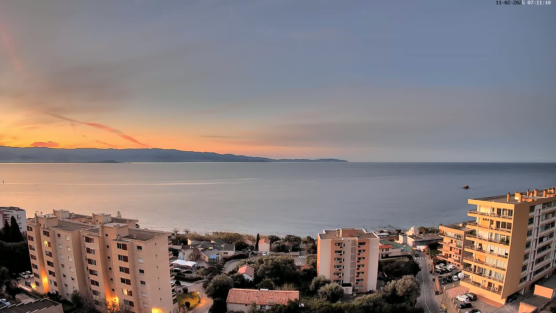 A coastal sunset scene shows several apartment buildings overlooking a calm sea and distant mountains under a mostly clear sky.