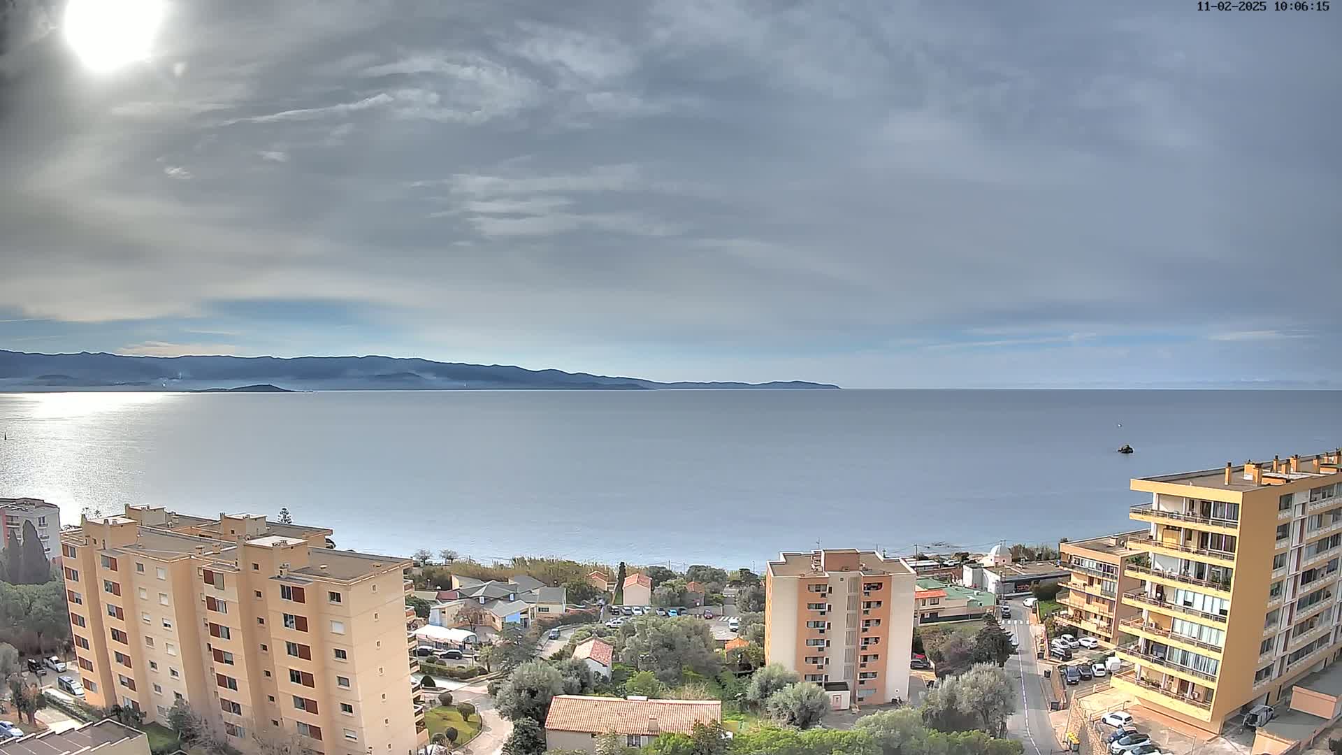 A partly cloudy day overlooks a calm sea with distant mountains, several apartment buildings, and green vegetation.