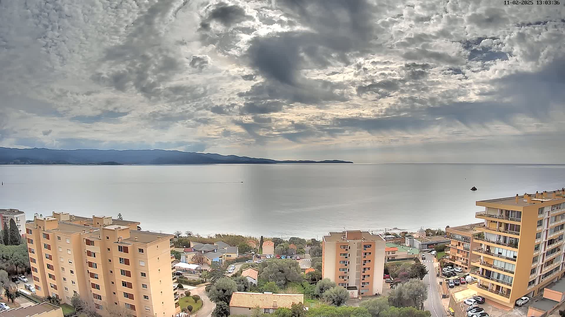 A coastal town with several apartment buildings overlooks a calm sea under a cloudy sky, with mountains visible in the distance.
