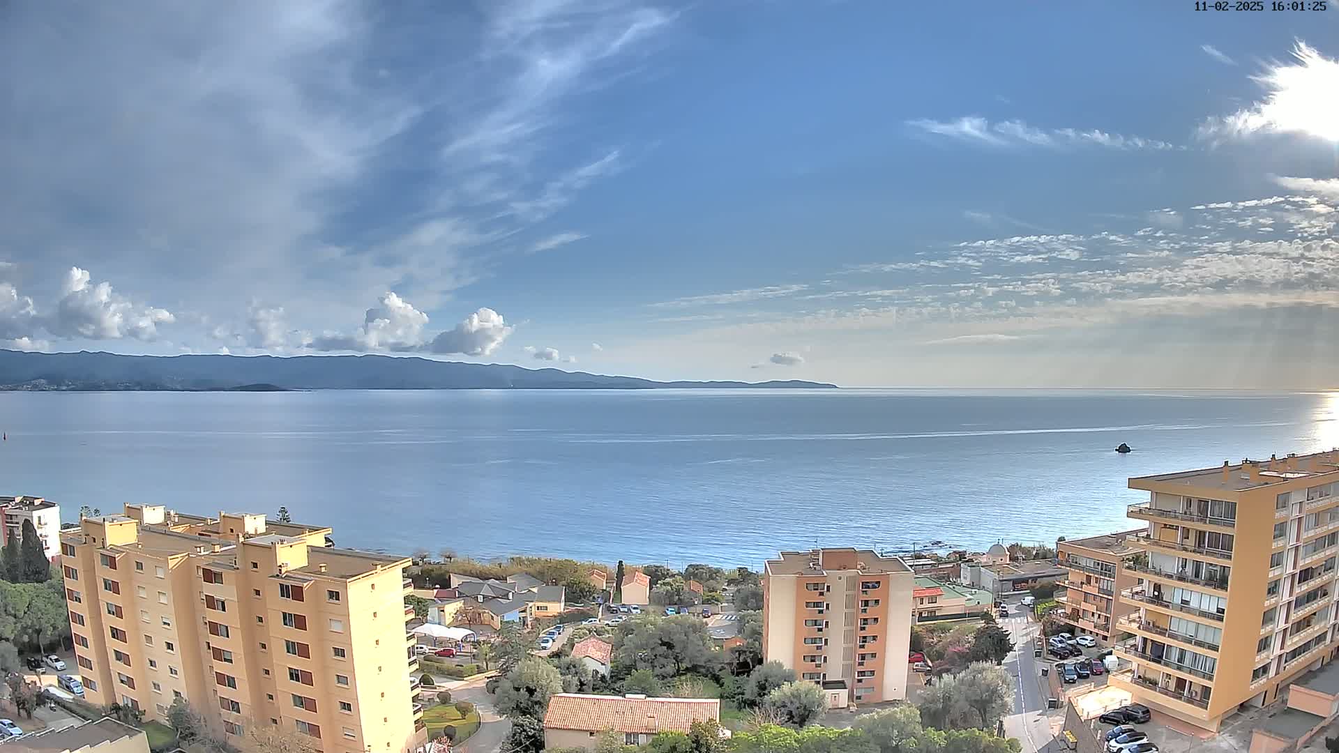 A partly cloudy sky overlooks a calm sea with distant mountains, and several apartment buildings are situated on a hillside overlooking the water.