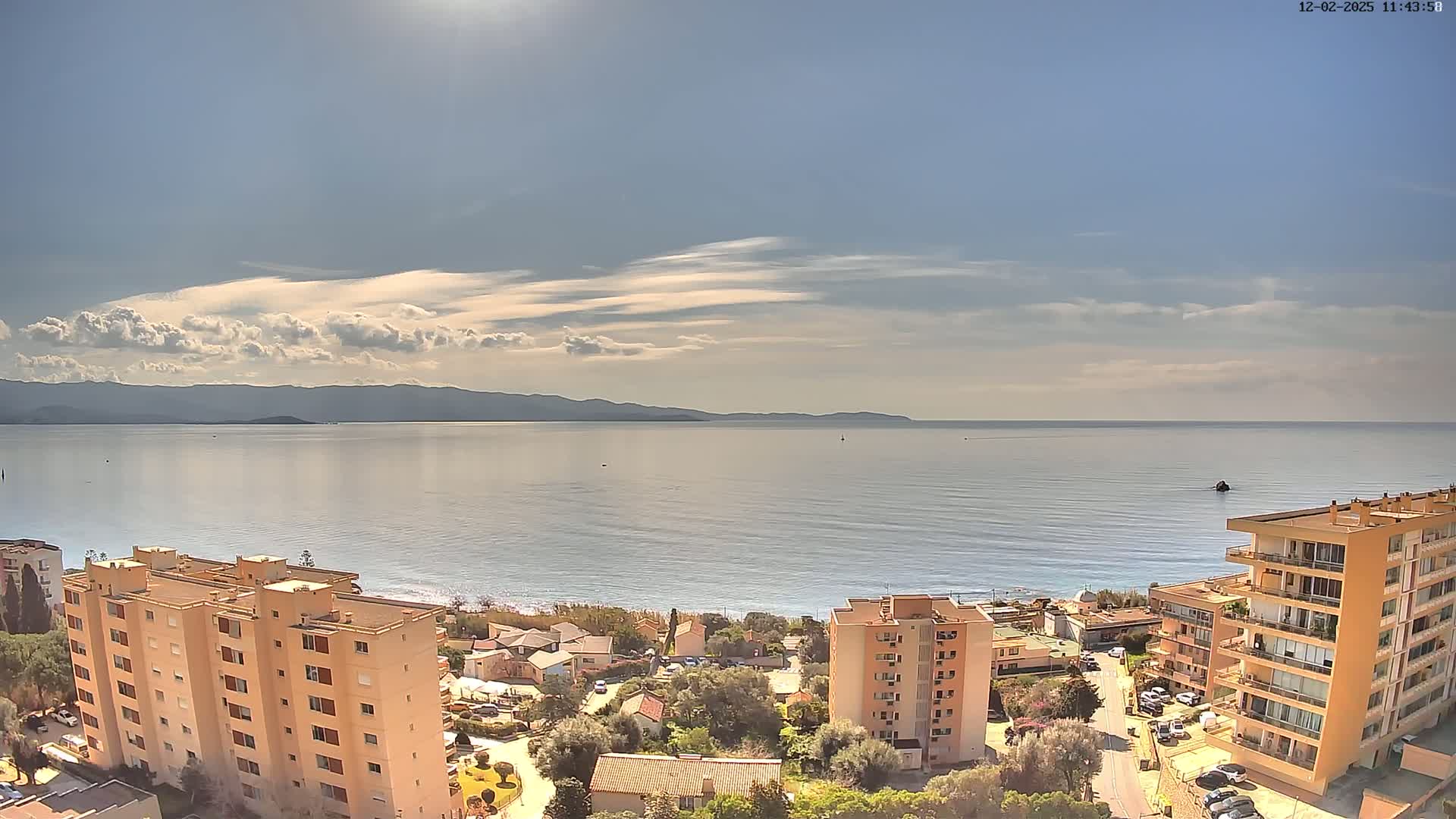 A partly cloudy day reveals a calm sea with distant mountains, overlooked by several apartment buildings and houses nestled near a beach.