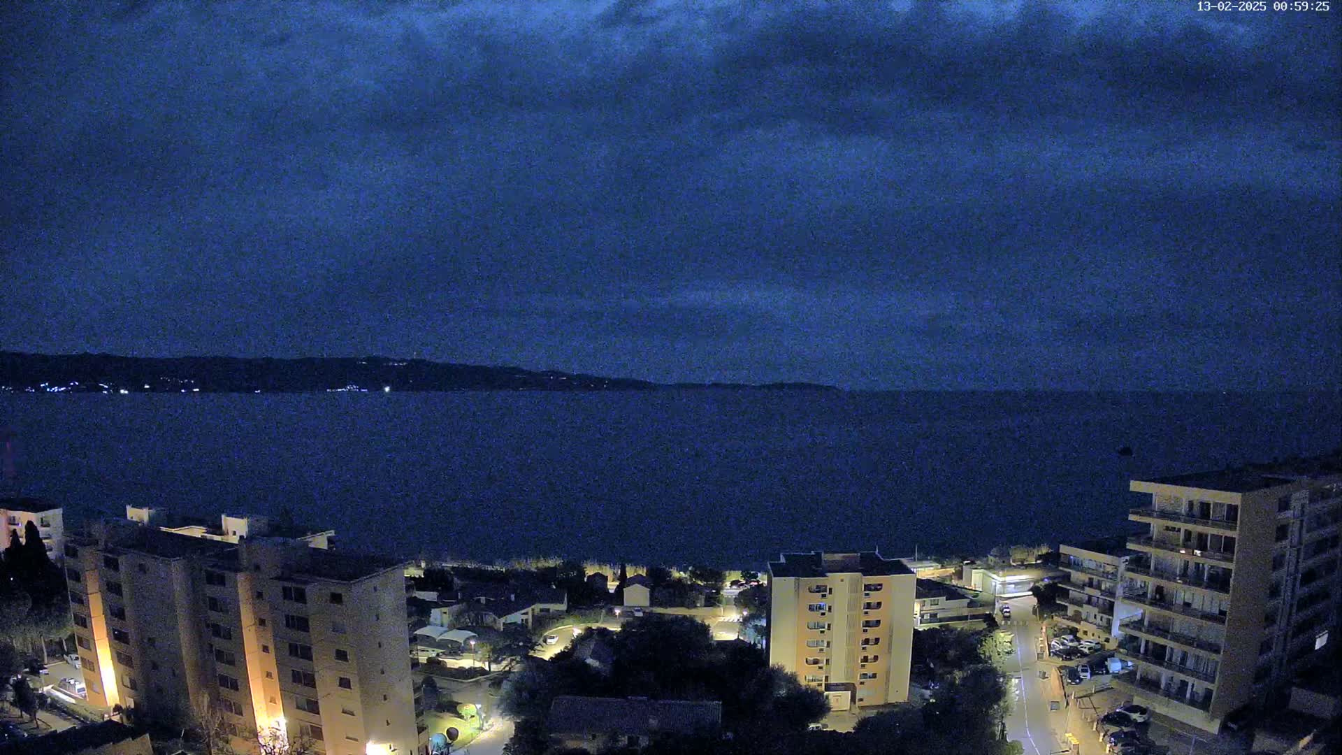 Nighttime view of a dark-blue sea under a cloudy sky, with several illuminated apartment buildings situated near a shoreline.