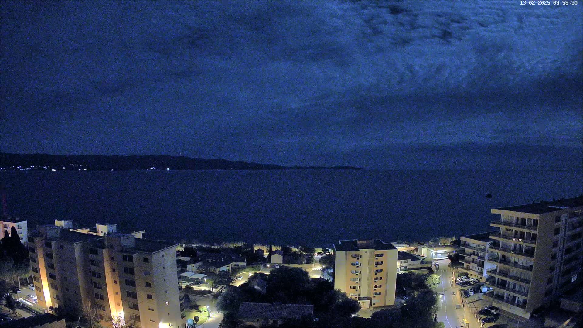 A nighttime view of a calm body of water under a dark, cloudy sky, with illuminated buildings and streets visible along the shoreline.