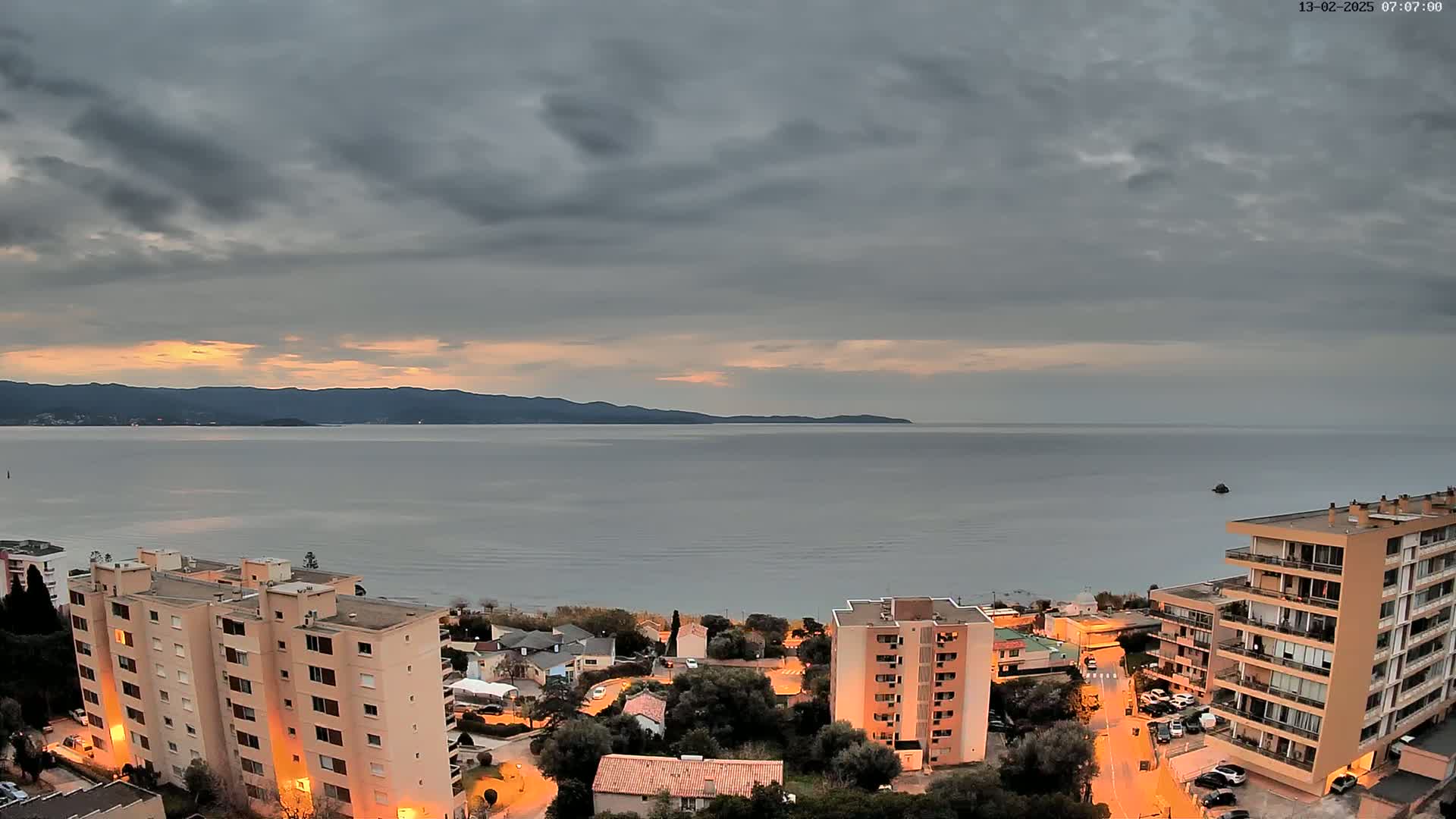A calm sea stretches to a distant mountain range under a mostly cloudy, dusky sky, with apartment buildings and streetlights illuminating a coastal town at sunset.