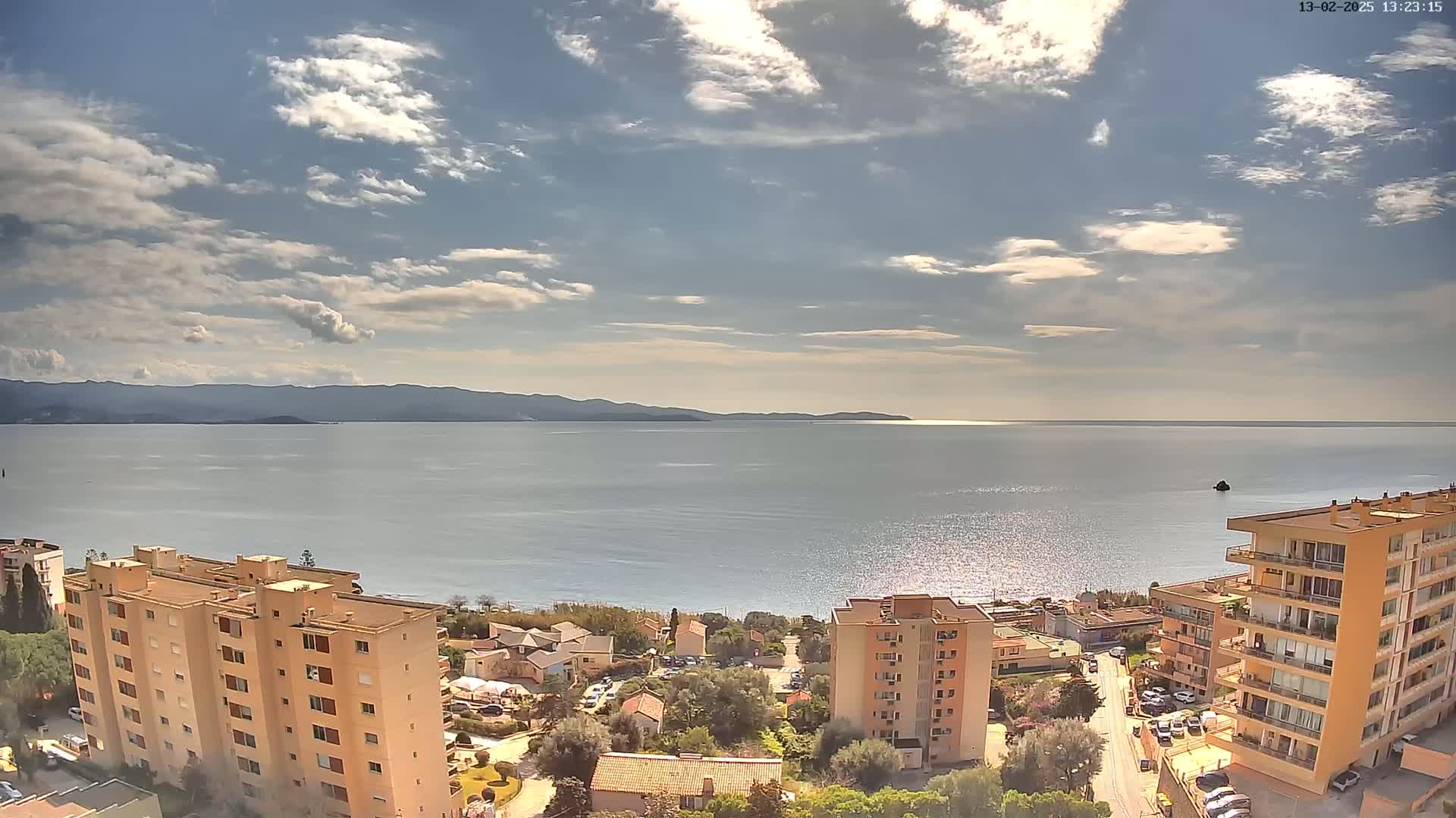 A partly sunny day overlooks a calm sea, distant mountains, and several multi-story buildings along a shoreline.