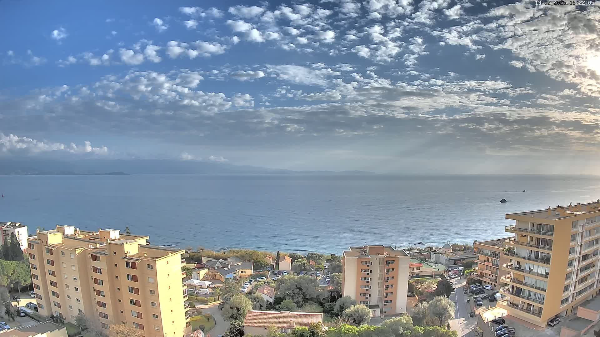 A coastal cityscape under a partly cloudy sky overlooks a calm sea with distant mountains.