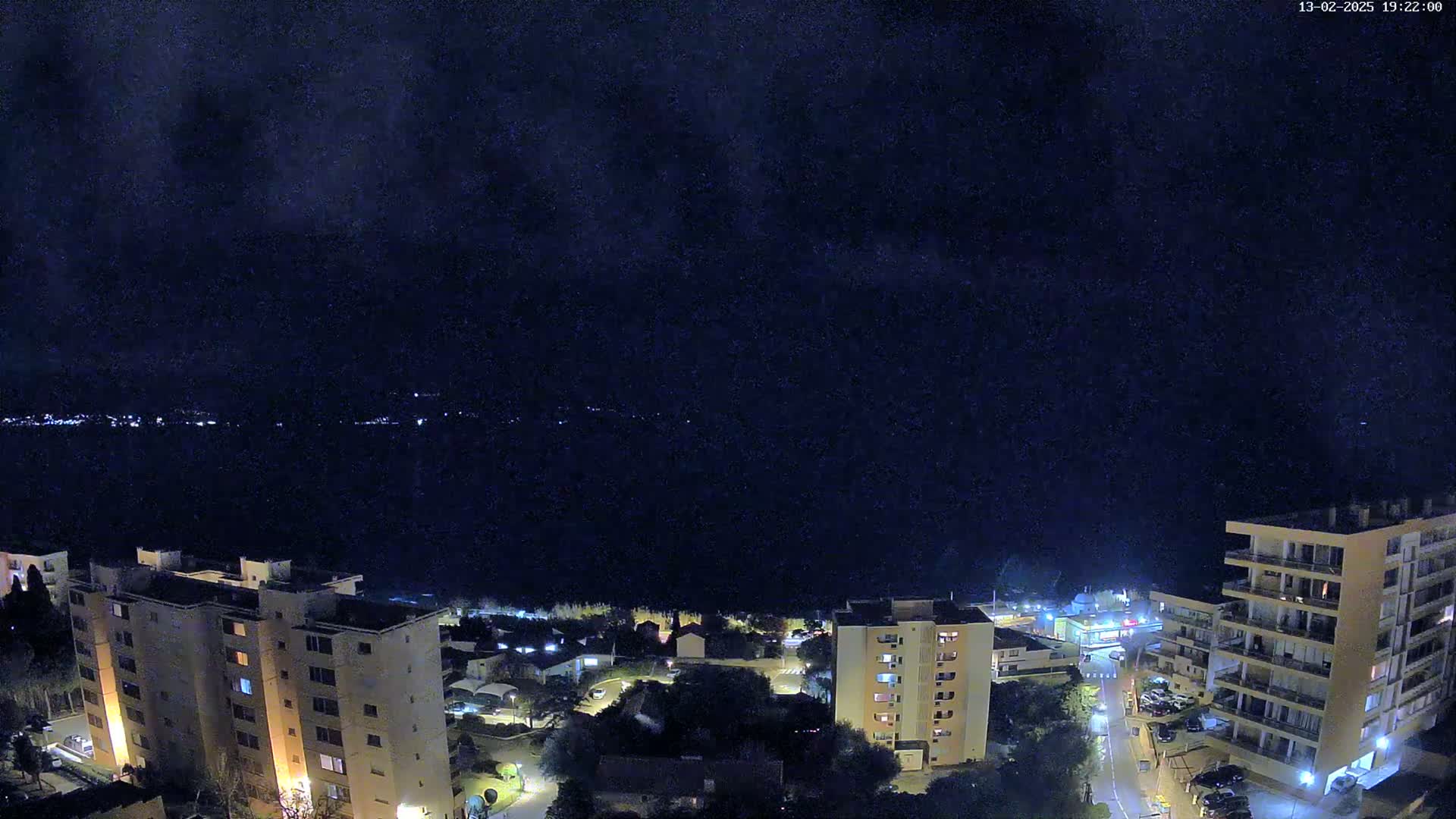 A nighttime, high-angle view shows a coastal town with several apartment buildings, a dark body of water, and a faintly visible line of lights across the water under a dark, mostly clear sky.