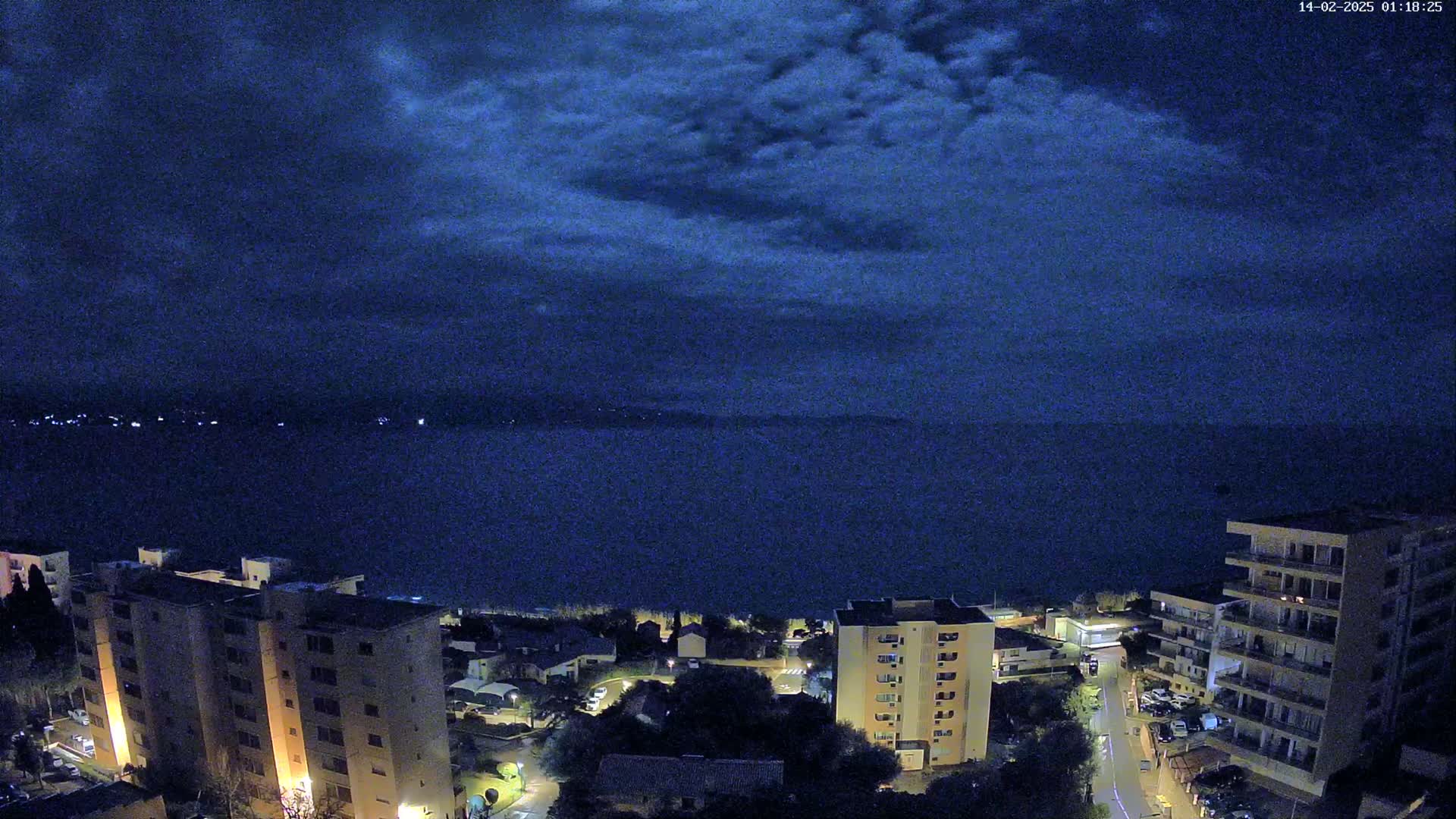 A nighttime, high-angle view shows a dark body of water under a cloudy sky, with several multi-story buildings and surrounding streets illuminated below.