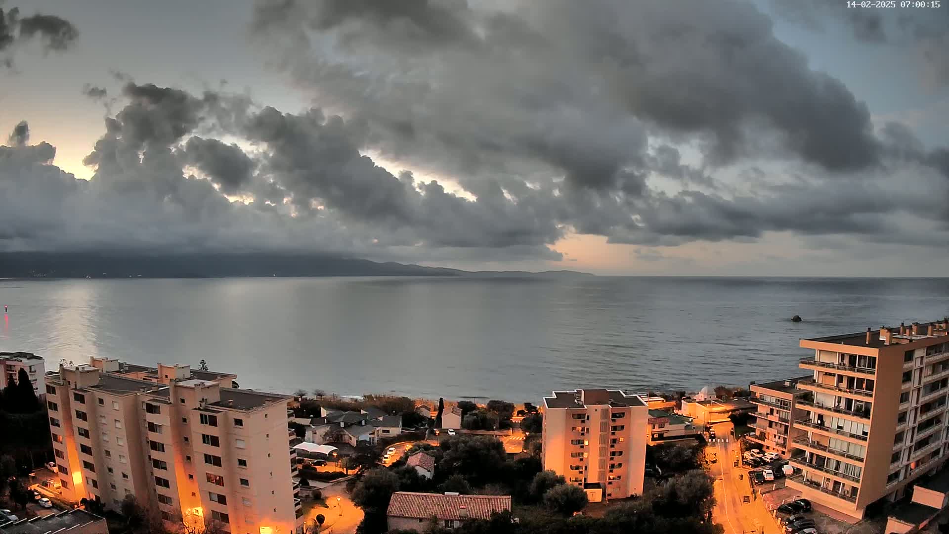 A coastal town with apartment buildings is illuminated at twilight under a cloudy sky, overlooking a calm sea and distant mountains.