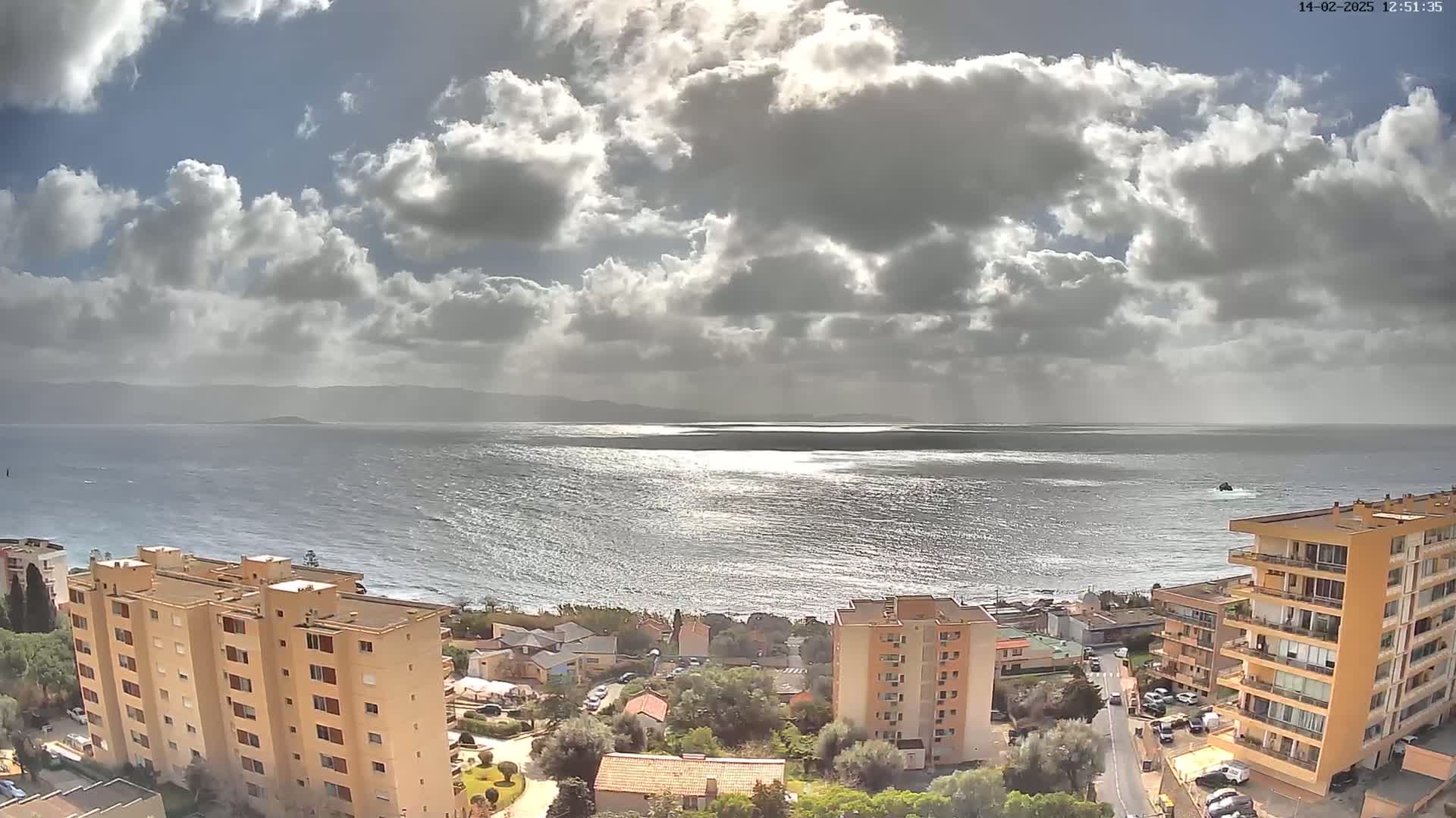 A partly cloudy day overlooks a calm sea with sun glinting on the water, from a high vantage point showing several multi-story buildings and a coastal town.