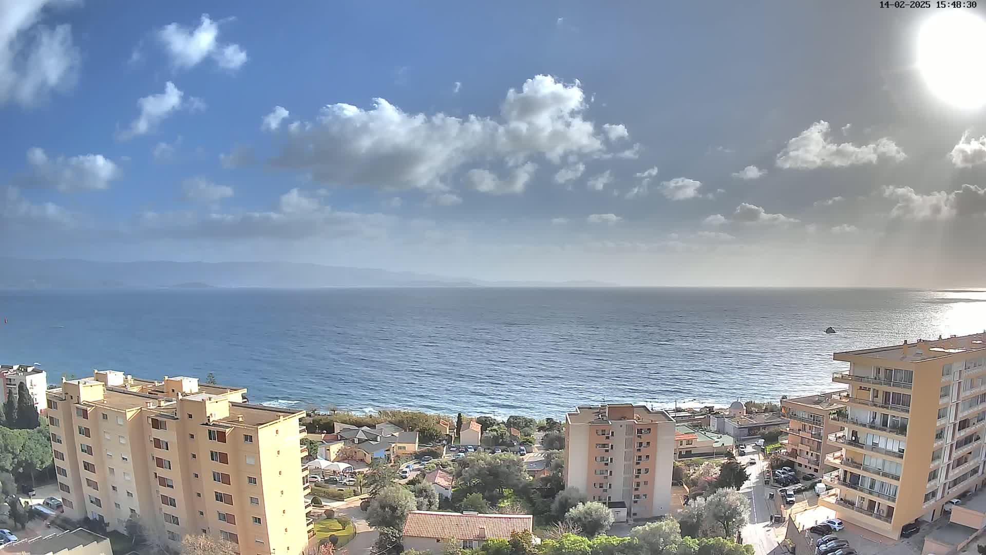 A partly cloudy sky overlooks a calm ocean with a shoreline of buildings and vegetation.