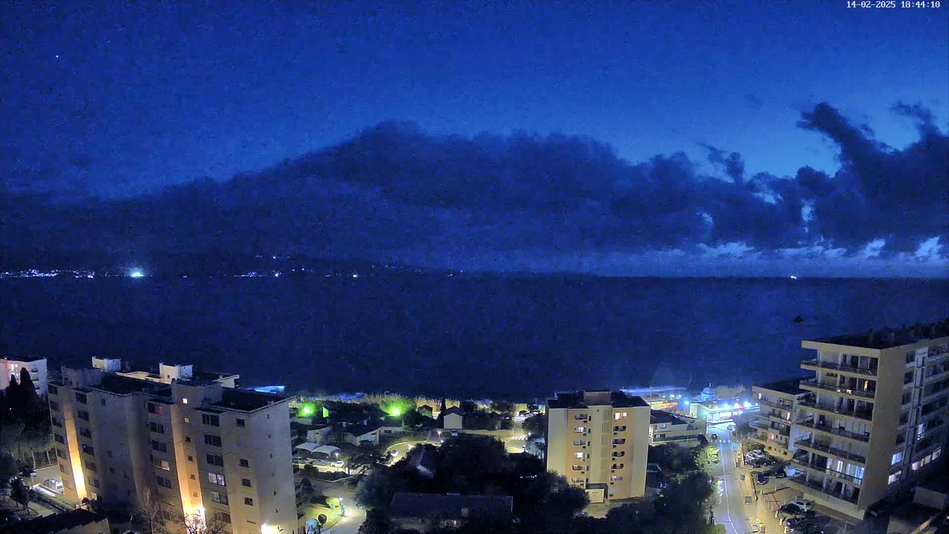 A nighttime view of a coastal town under a dark blue sky with scattered clouds, showing apartment buildings and a dark ocean stretching to a distant shoreline.