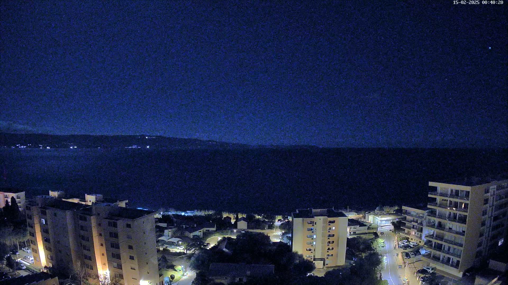A nighttime view of a coastal town with several apartment buildings overlooking a dark body of water under a clear, starlit sky.