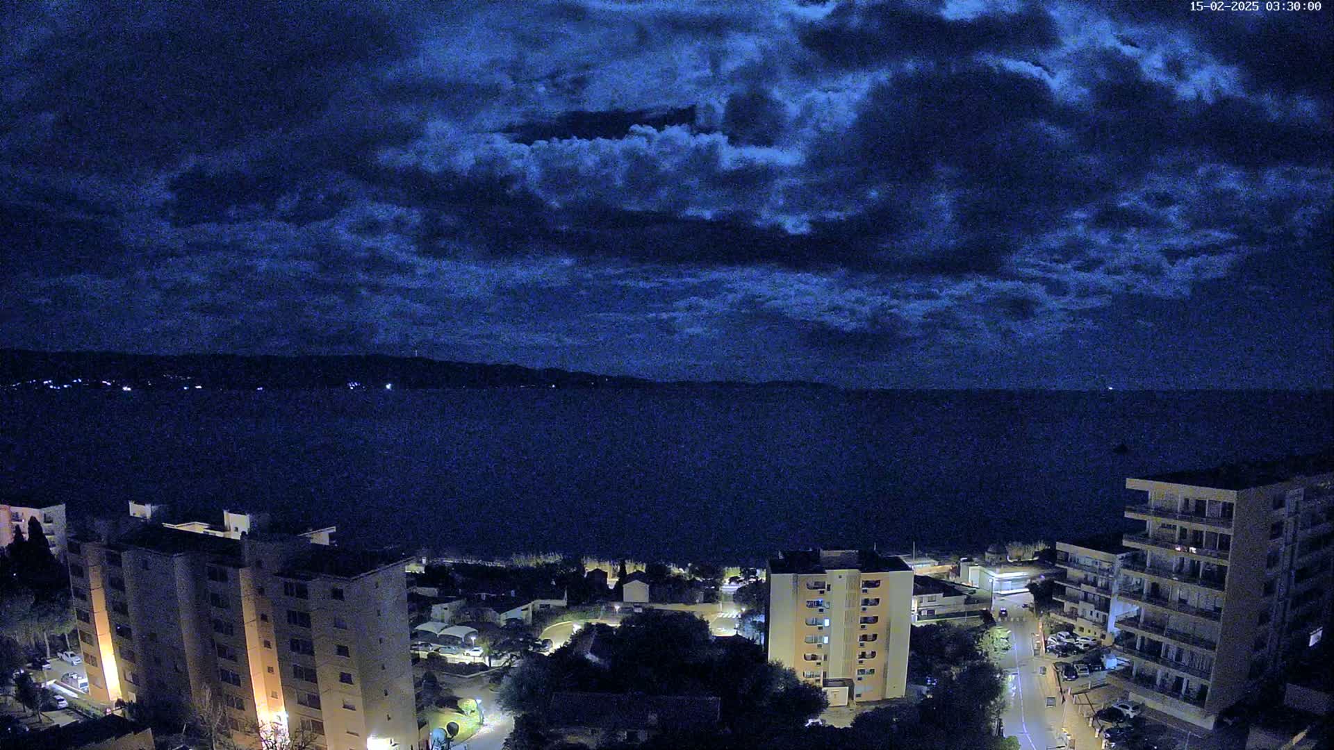 A nighttime coastal scene shows a dark, cloudy sky over a calm body of water, with lit buildings and streets visible along the shore.