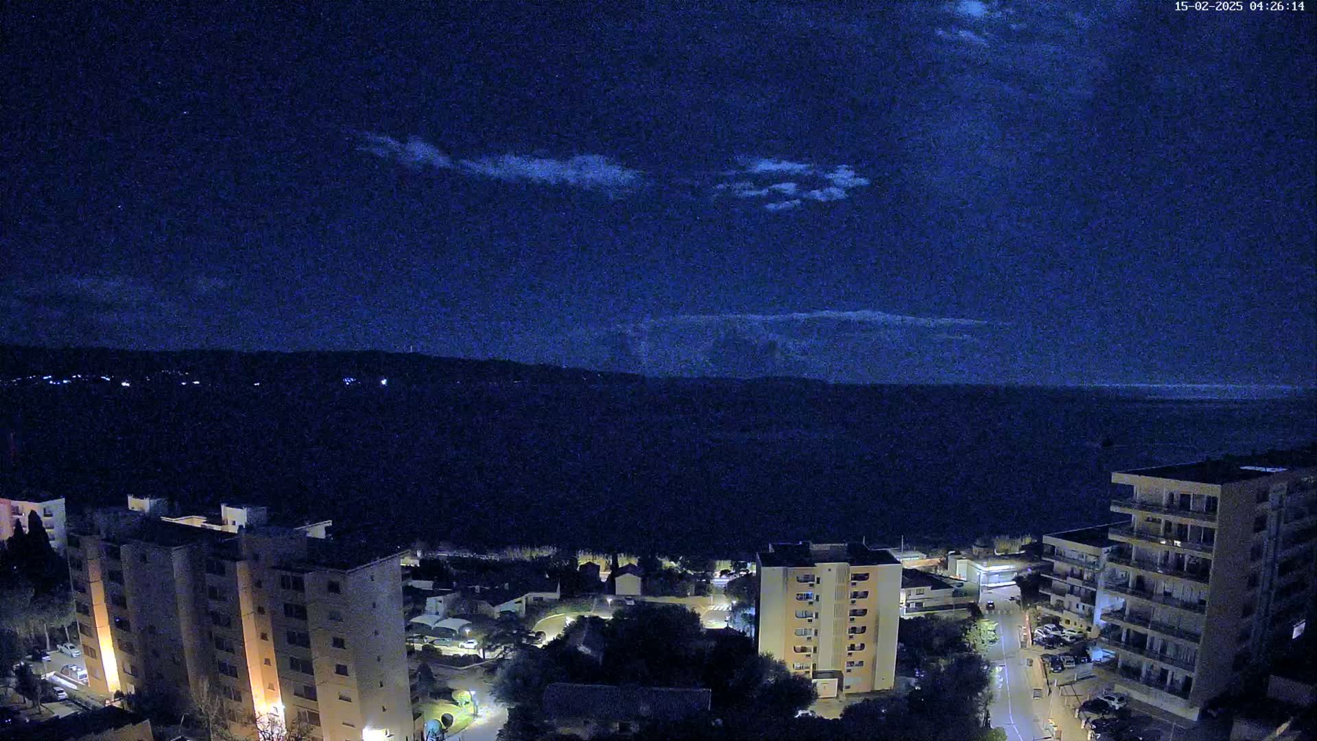 A nighttime aerial view of a coastal town with apartment buildings under a dark blue sky with a few clouds.