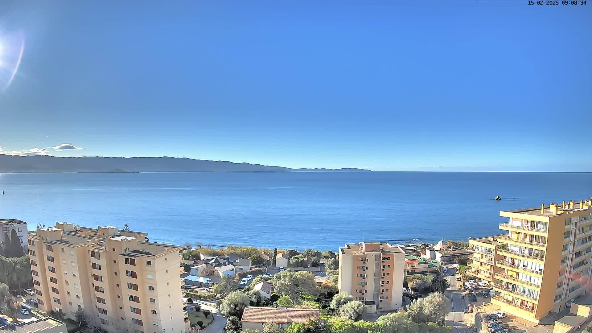 A clear, sunny day overlooks a calm blue sea with distant mountains and several apartment buildings in the foreground.